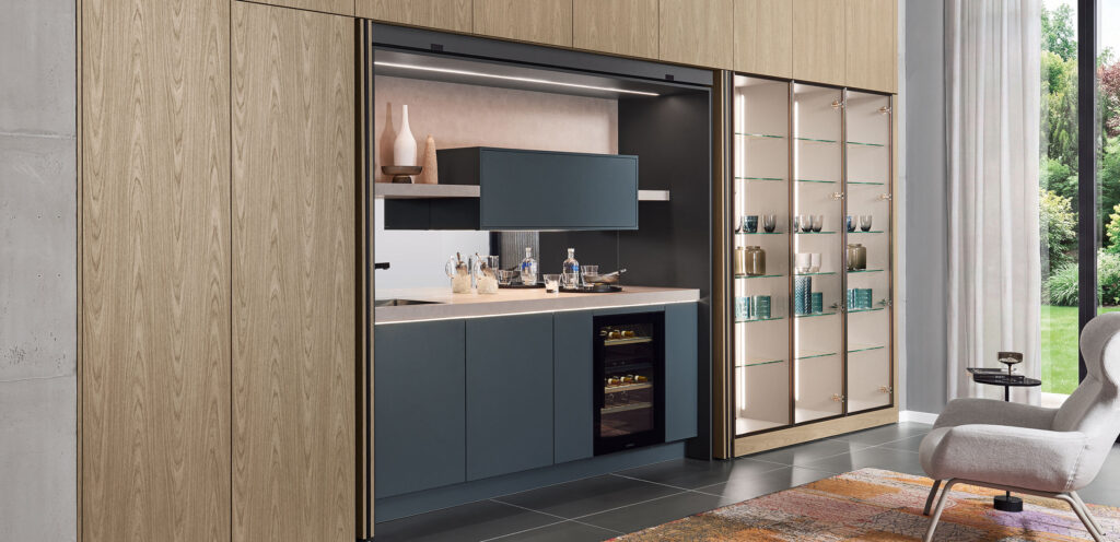 Geöffnete Pocket-Door-Schrankwand in Holzoptik mit blauer Nische, Weinkühlschrank und Glasvitrine