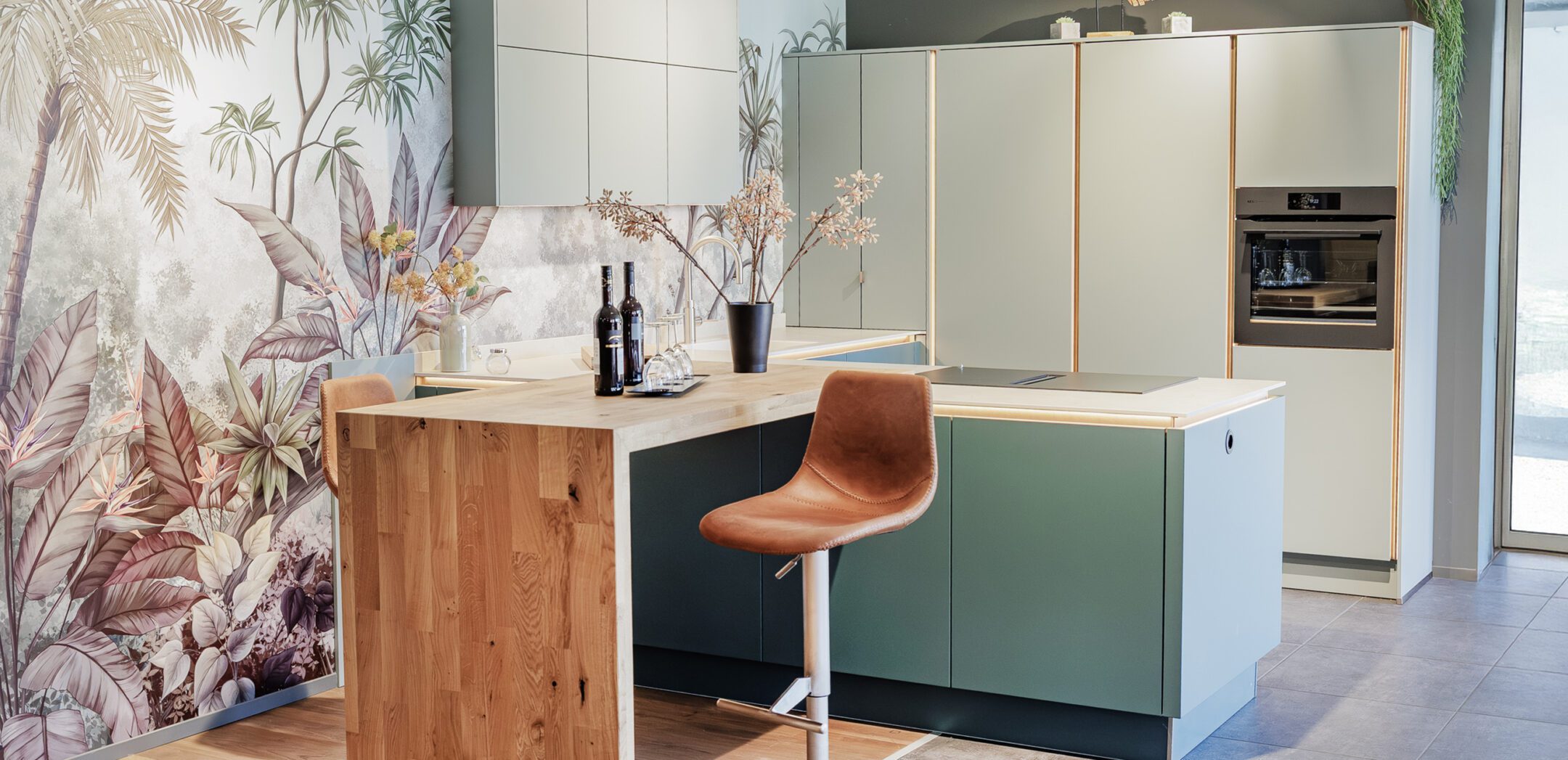Moderne grüne Inselküche mit Holztisch-Anbau und tropischer Motiv-Tapete.