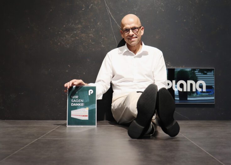 Rüdiger Schoeller sitzt entspannt am Boden vor einer dunklen Wand mit PLANA-Logo und hält das Focus-Money Dankesschild