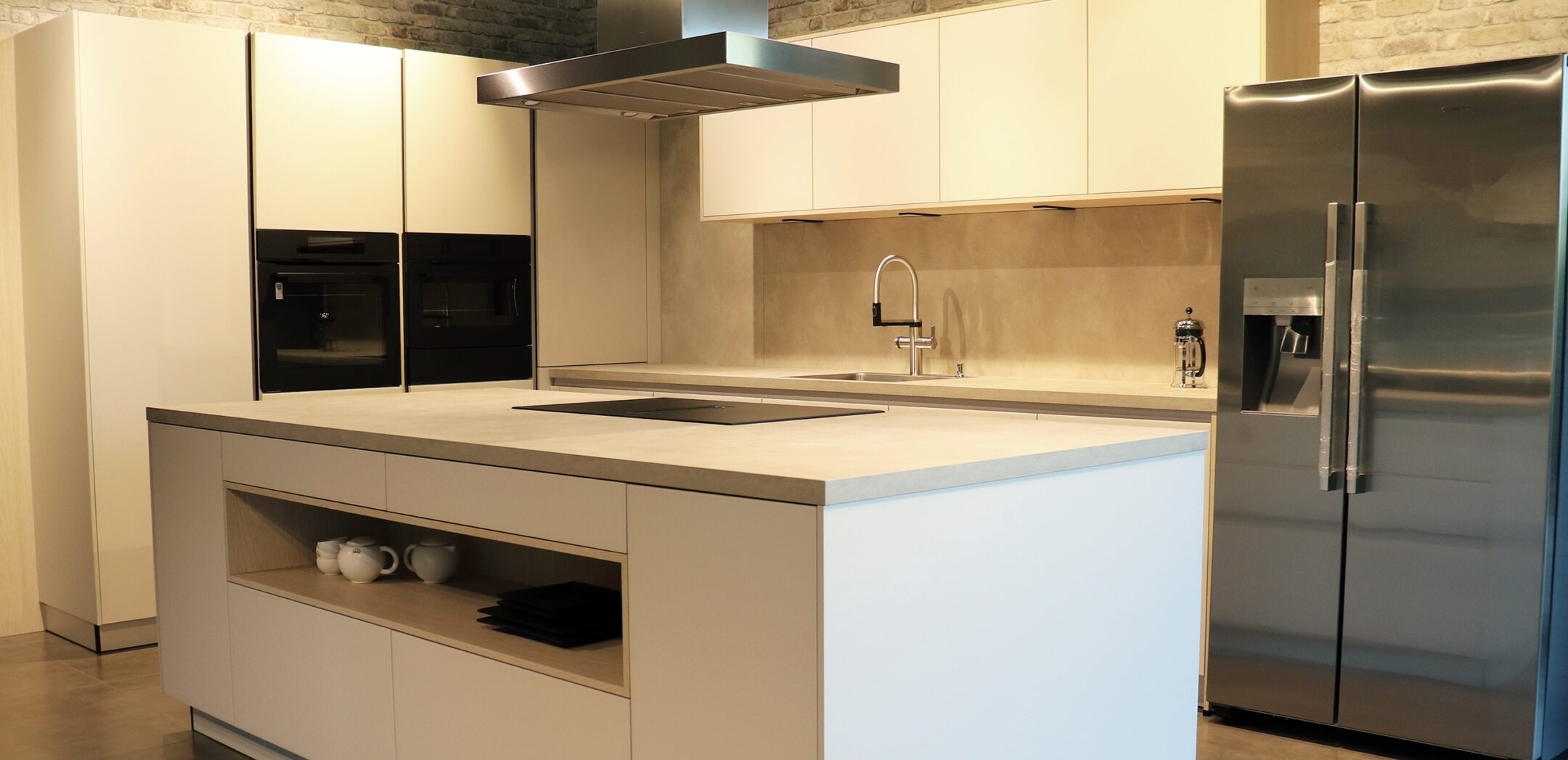 Große Kücheninsel in hellen Tönen mit integriertem Kochfeld und Side-by-Side Kühlschrank im Hintergrund.