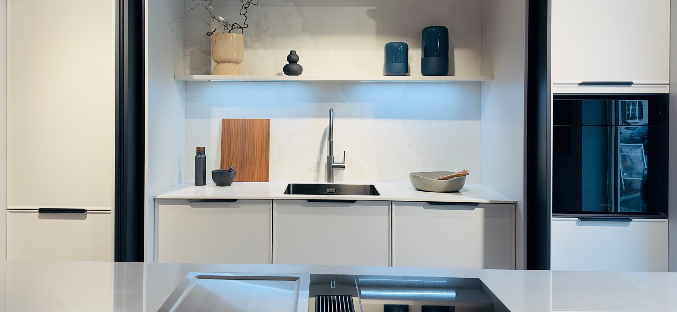 Helle Einbauküche mit weißer Arbeitsplatte, schwarzer Spüle und dekorativem Wandregal mit Beleuchtung.