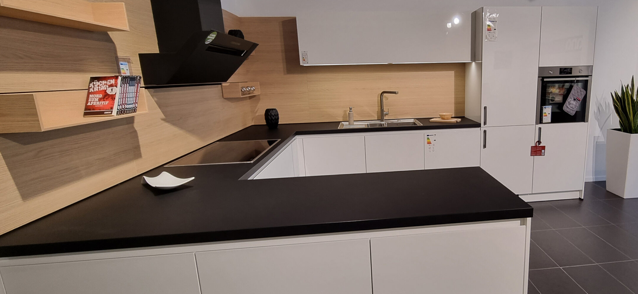 Weiße U-Küche mit schwarzer Arbeitsplatte und Holznischenrückwand im PLANA Küchenstudio Waldshut-Tiengen