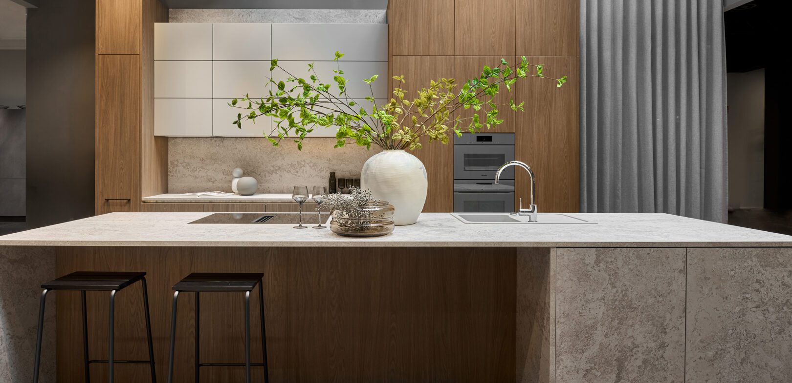 Moderne Wohnküche mit großer Kochinsel aus hellem Naturstein, eleganten Holzfronten, integrierten Küchengeräten und Barhockern.