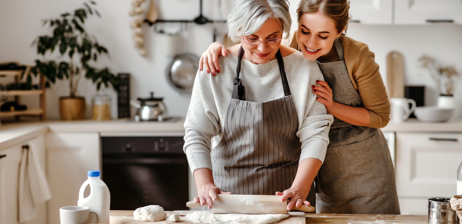 Ältere Dame mit Welholz rollt Teig aus, jüngere Frau umarmt von hinten
