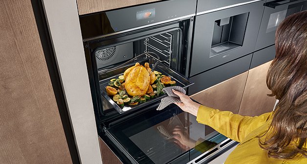 Frau holt ein Hähnchen aus dem Backofen, zwei weitere Siemens Einbaugeräte zu daneben