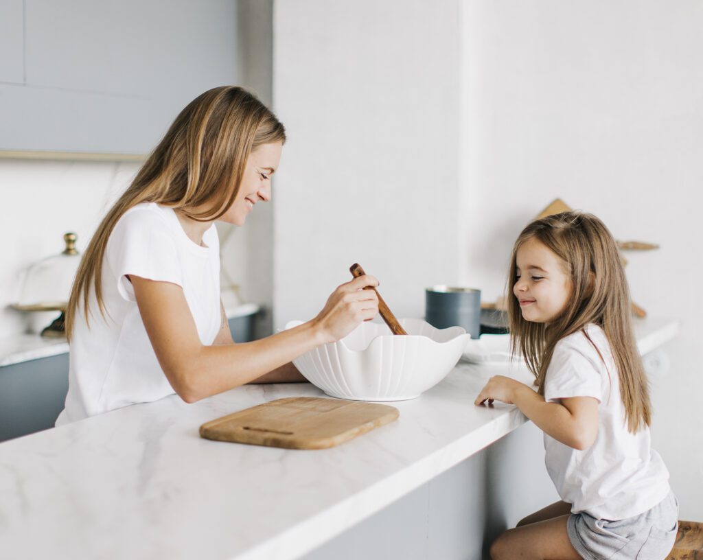 Mutter und Tochter backen gemeinsam in einer hellen, modernen Küche an einer weißen Marmor-Theke.