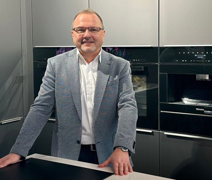 Ein Küchenplaner im blauen Sakko lehnt an einer hellen Kücheninsel.