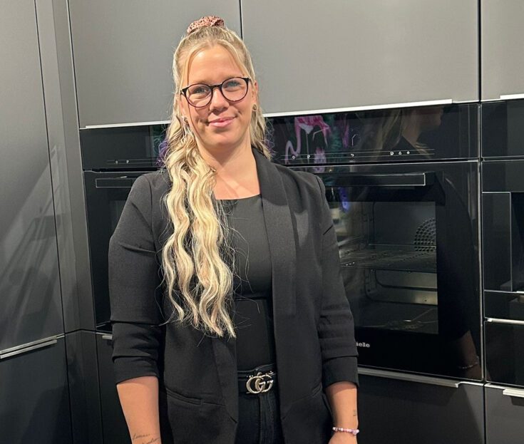 Eine Küchenberaterin mit Brille lächelt vor einer dunklen Schrankwand mit Einbaugeräten.