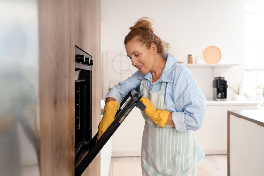 Eine lächelnde Frau mit hochgesteckten Haaren und gelben Gummihandschuhen öffnet die Tür eines modernen Einbaubackofens in einer hellen Küche.