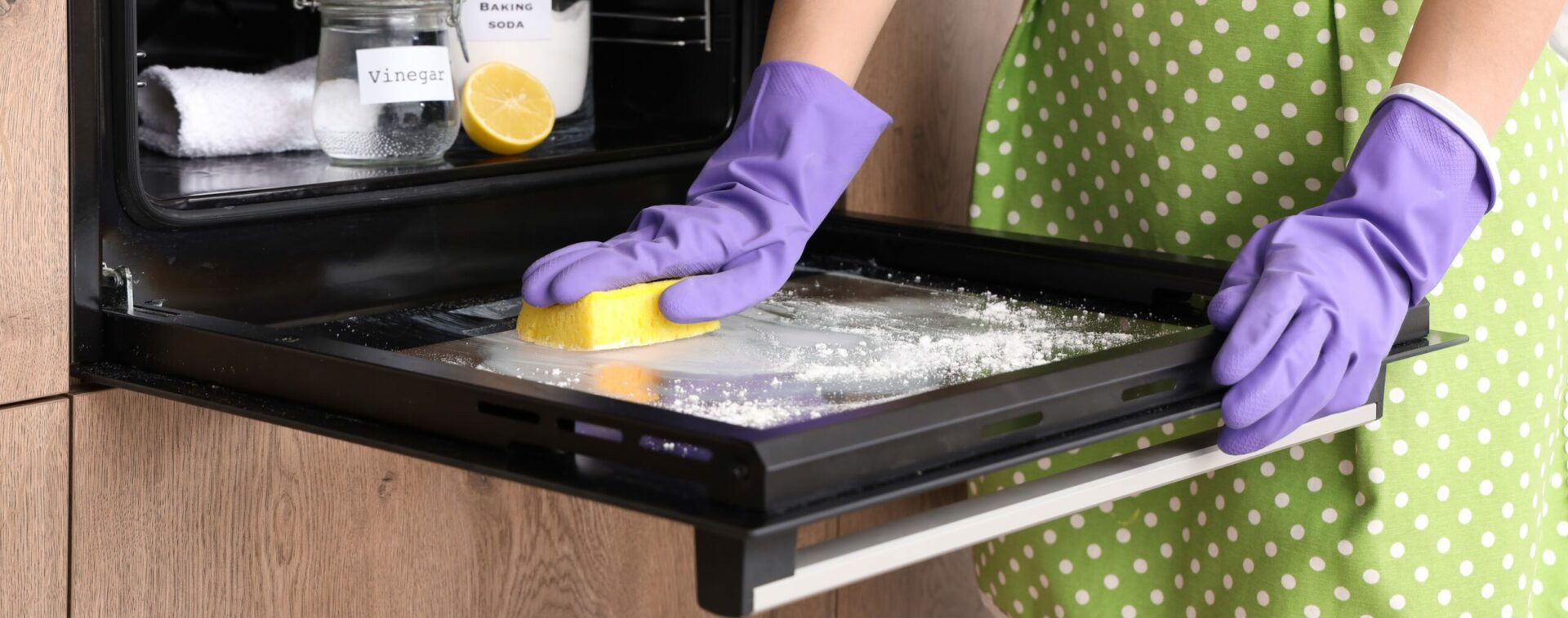 Eine Person in lila Handschuhen schrubbt mit einem gelben Schwamm und weißem Pulver die Innenseite einer offenen Backofentür