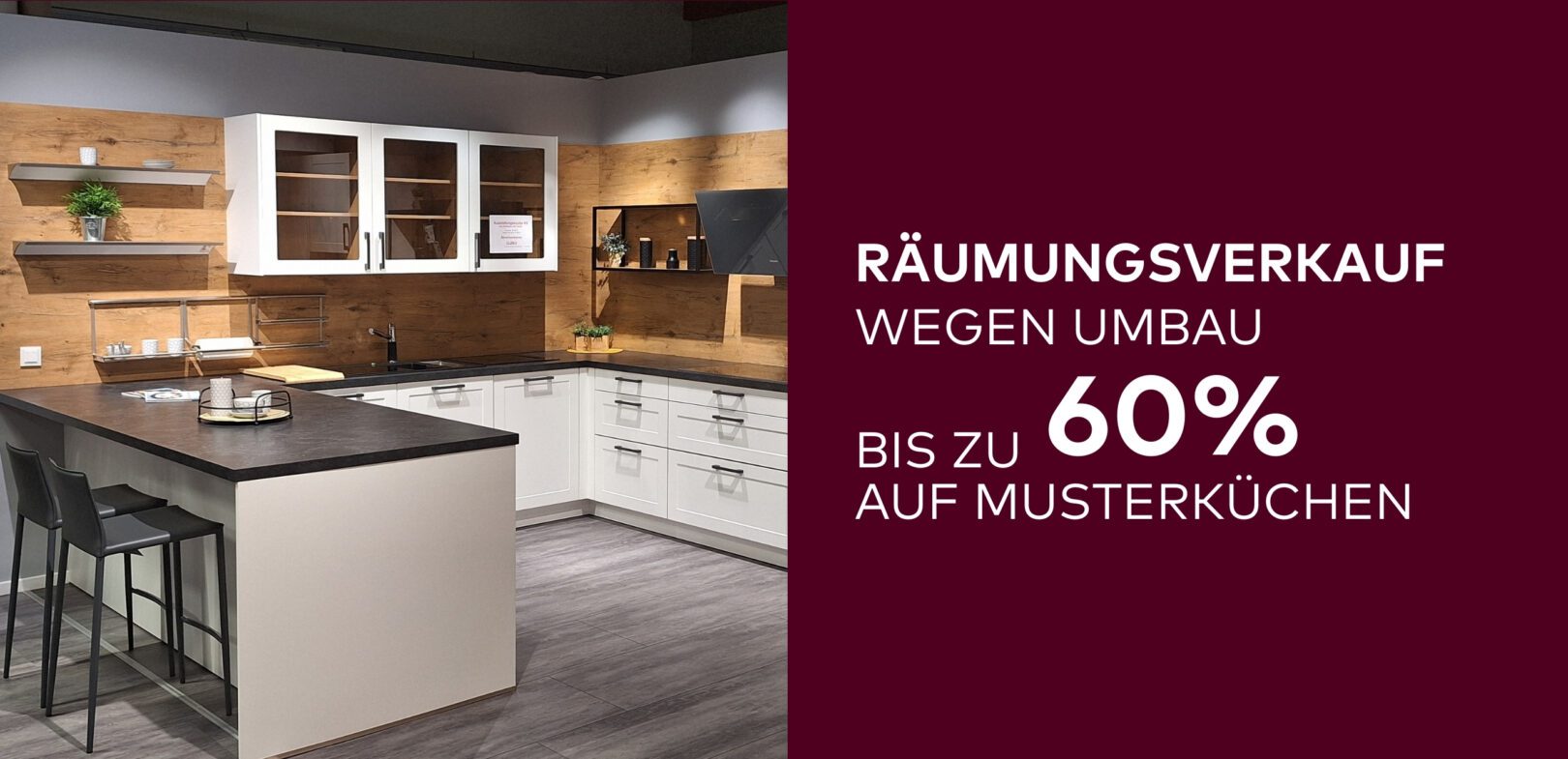 Zweigeteiltes Bild: Die linke Seite zeigt eine ausgestellte Einbauküche im Landhaus-Stil mit Holzwänden, weißen Schränken, dunkler Arbeitsplatte, einer integrierten Bar und Barhockern. Die rechte Seite ist ein dunkelroter Textbereich mit der weißen Überschrift: 'WIR BAUEN UM!' und der großen Zahl '60%' darunter. Der restliche Text lautet: 'BIS ZU AUF MUSTERKÜCHEN'.