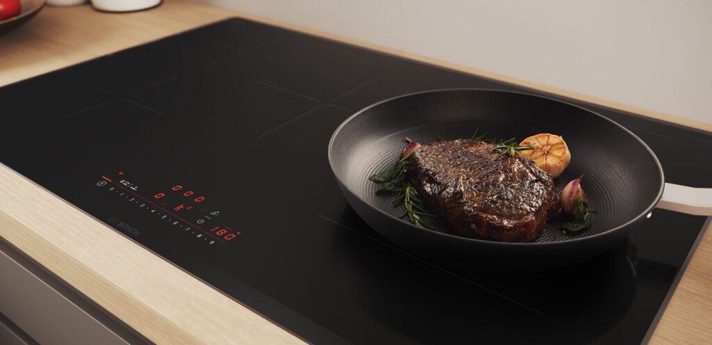 Modernes Bosch Induktionskochfeld mit roter Touch-Bedienung und einer Bratpfanne, in der ein saftiges Steak mit Knoblauch und Kräutern brät.